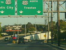 A multilane divided highway approaching an interchange with a sign over the road reading U.S. Route 130 north Trenton with two down arrows