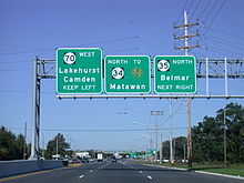 A six-lane divided highway approaching an intersection with a set of three green signs over the roadway. The left sign reads Route 70 west Lakehurst Camden keep left, the middle sign reads north Route 34 to Garden State Parkway Matawan, and the right sign reads Route 35 north Belmar next right.