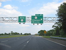 A four lane freeway at an interchange. Two green overhead signs stand over the road with the left one reading south Route 55 and the right one reading exit 27 Route 47 Vineland Millville with an arrow pointing to the upper right