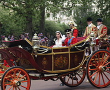 Couple sitting in a decorated horse-drawn open-top carriage, with two footmen in livery sitting behind the newly-weds.