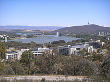 A group of multi-story office buildings. A lake, mountains and a jet of water are visible in the background.