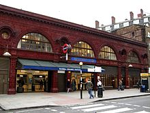A red glazed terracotta building. The first storey above ground features four wide, storey-height semi-circular windows with smaller circular windows between above which is a dentil cornice. Below the two right-most windows, the station name, "Russell Square Station", is displayed in gold lettering moulded into the terracotta panels. A blue tiled panel above the entrance says "Underground".