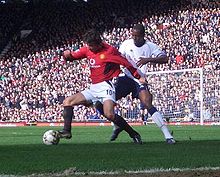 A photograph of two men playing football. The man on the left, who is wearing a red shirt, white shorts and black socks, is shielding the ball from the man on the right, who is wearing a white shirt, navy blue shorts and white socks with navy blue trim.