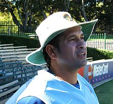 A man with dark skin in a light blue sleeveless pullover and dark blue t-shirt facing to the right.  He is wearing a wide-brimmed white hat and is standing in front of some empty bleachers with trees further behind.
