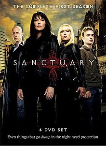 A DVD boxset cover consisting of four people standing behind a logo saying "Sanctuary". In the front is a middle-aged brunette woman wearing black. To her right standing behind her is a bald man with a black leather overcoat. To her left is a young blonde woman, also sporting a black leather coat and a man with curly hair. In the background, modern skyscrapers can be found on the left of the set, while a gothic cathedral can be found on the right.