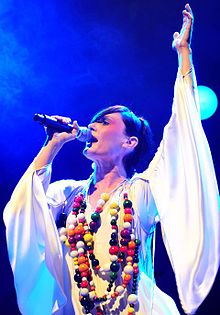 A woman singing into microphone, looking forward, left arm is raised in a white robe with a multicolor necklace.