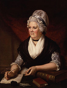 Half-length portrait of a elderly woman sitting at a desk surrounded by books and papers and holding a quill.