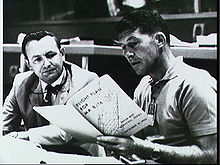 Two men seated at a desk, both reading from a large book the younger man is holding