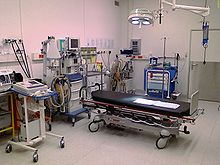 Color photograph of a room designed to handle victims of major trauma. Visible are an anesthesia machine, a Doppler ultrasound device, a defibrillator, a suction device, a gurney, and several carts for storing surgical instruments and disposable supplies.