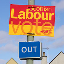 A yellow, orange and red election sign reading "Scottish Labour: Vote Scottish Labour" attached to a lamppost above a road sign stating "Out"
