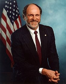 Head and shoulders of a man in his mid-fifties, seated with hands clasped. He is Caucasian and wears glasses and a black suit with a white dress shirt and a red tie. His head is balding and he has a gray beard with traces of brown hair. A U.S. flag is in the background.