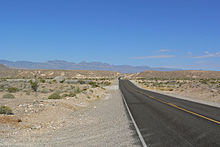 A two-lane asphalt highway passes through a desert landscape dotted with sagebrush and cacti as it heads towards the distant mountains.