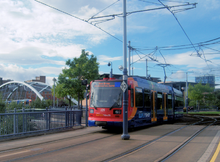 A Sheffield Supertram in current blue, orange and red Stagecoach livery. The Tram shown is cross the Park Square junction. Behind it is the bridge connecting the junction and tracks to Sheffield city centre. The whole square (now 20 years since redevelopment) is covered in trees and plants and new buildings can be seen beyond them.