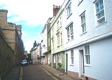A narrow road, pavements either side; on the left, a stone wall; on the right, a row of terraced three storey houses, the upper storeys overhanging the ground floor