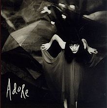 A black-and-white photo of a Caucasian woman leaning forward while holding the ends of a flowing black dress. In the corner, "Adore" is displayed in white handwriting.
