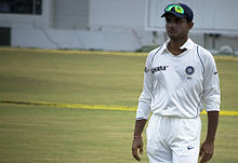 An Asian cricketer in cricket whites, wearing a dark blue baseball cap, with sunglasses on top.  He is standing on his own on a cricket pitch.