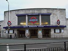 A curved white stone building with three glazed screens above a low three part entrance. The central screen features the London Underground logo in coloured glass (a red circle crossed by a horizontal blue bar carrying the UNDERGROUND name)