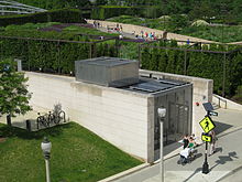 A small rectangular building with two walls of buff colored stone and two walls of glass. Solar panels are visible on the roof, and a "P" (for parking) sign is on a stone wall. Pedestrians are in front of a garden is behind the structure.