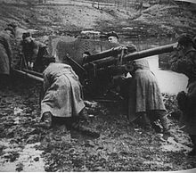 A large gun trapped in mud. Several men in long, heavy coats are pushing on it trying to get it free.