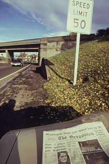 A sign next to a highway says "Speed Limit 50". A newspaper in the foreground has an article about the new speed limit.