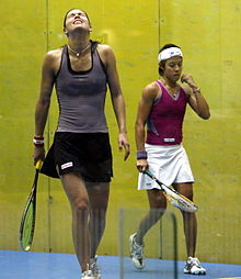 A female squash player in a bluish top throws back her head in annoyance while another squash player in a purple top and a white headband walks by
