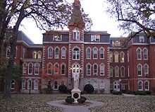 A statue sits in front of a large four-story red brick building. To the right of the entrance, the building goes in a little ways, and then back out. To the left of the entrance is a small two-story area connecting the main part of the building to the rest of it. Above and behind the two-story part is a four-story part of the building