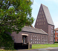 An angular taupe-coloured church on a bright sunny day. The church fills the image with the exception of a flat piece of green grass and grey pavement at the church's entrance and the dark green silhouette of trees along the left of the picture. The single-storey church has many small slitted triangular and rectangular windows across its façade, except for a dark rectangular entrance in which a man stands. At the far end of the church is a triangular spire standing tall and bold against a light blue sky at around six or seven times the height of a man.