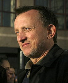 Headshot of a middle aged man looking to his right. The man has short hair and light stubble. He appears to be smiling slightly, and is wearing a dark coat.