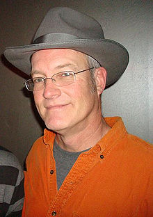 A middle-aged Caucasian man wearing spectacles, a gray fedora and an orange shirt smiles for a camera.
