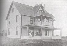 A three story house with four men and two children on the front porch and three women on the second floor balcony