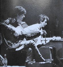 Black-and-white picture of two young men playing electric guitars, the guitarist in the foreground wearing a leather jacket and the one in the background a white collared shirt. Other individuals are visible in the background