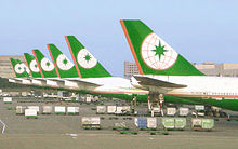 Row of eight aircraft tails in identical livery, lined at airport terminal, surrounded by cargo and equipment.