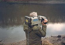 A man standing beside a river holding a camera with the Tyne Tees logo.