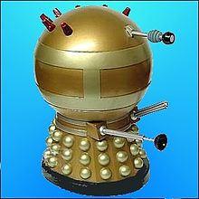 Full length image of a squat Dalek, painted overall in gold. It has brass-coloured hemispheres, collars and two wide bands around the spherical head section, the upper rear portion of which is adorned with seven red lights.