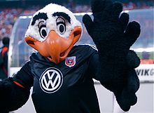 A black and white costumed bald eagle mascot with exaggerated features and an orange beak raising his wings. He wears a black soccer jersey with a white WV logo and the team's shield on it.