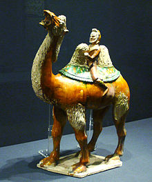 A glazed figurine of a red camel, which is being rided by a bearded merchant in green clothing