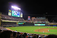 Baseball field at night, scoreboard displays a player, Twins "M" and "Stp" decorate a large neon sign