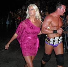 A Caucasian woman in a dark pink dress walks arm in arm with a Caucasian male wearing purple tights and a jewel-encrusted belt.