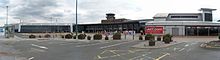 The frontage, consisting mainly three rows of glass panes, of a long building. Above the glass is a concrete section containing the words "Leeds Bradford International Airport" in capitals and, to the right, an aeroplane symbol. Beyond this, the concrete section continues diagonally downwards to the ground. To the left of this support is an entrance which cuts into six panes of the two lower rows of glass panels, and some passengers pulling suitcases are entering the building. Two smaller entrances to the left replace four of the ground-level panels. In front of these are railings. In the foreground are roads, pedestrian walkways, bollards and five large free-standing box-shaped objects.