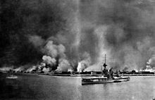 A city burns in flames; in the foreground, a large battleship sits in the water.  A smaller vessel is berthed next to her.  Another small ship sails away from the city on the left.