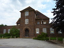 A photograph of a large derelict brick building from the 1930s. The front of the building has a large rounded archway in art deco style. The lower windows are boarded up and the panes of many of the upper windows are smashed