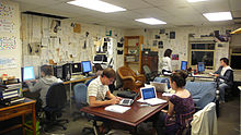 Several young men and women sit in office chairs working at computers around a room where the walls are covered in printed pages. A central wood table and bookcases are featured.