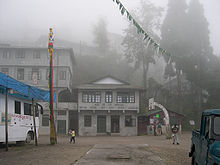 A two-storeyed building to the right of a four-storeyed building. A basketball court in the foreground and a couple of vehicles parked on either side. Foggy atmosphere, with trees in the background.