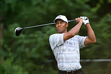 Tiger Woods, an African-American male in his early-30s, looks serious and wears a white hat and blue greyish polo shirt. On both his hands, he holds a golf club, after swinging it, and sports a white glove on his left hand.