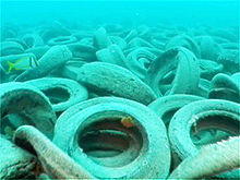 An endless bed of old, skummy tires rests piled upon the ocean's floor; a small yellow fish swims by the left of the photo.