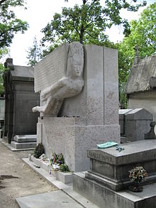 A large, rectangular, granite tomb. A large, stylised angel, leaning forward is carved into the top half of the front. There are a few flowers beside a small plaque a the base. The front is covered in red daubings and graffiti.