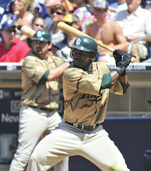A man wearing a camouflage baseball jersey, white pants, and a green batting helmet holds a baseball bat, ready to swing