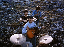 The Beatles performing music in a field. In the foreground, the drums are played by Starr (only the top of his head is visible). Beyond him, the other three stand in a column with their guitars. In the rear, Harrison, head down, strikes a chord. In the front, Lennon smiles and gives a little wave toward camera, holding his pick. Between them, McCartney is jocularly about to choke Lennon.