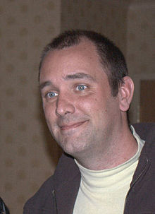 A brown-haired man wearing a lime shirt and brown jacket smiles and looks at something off-screen.