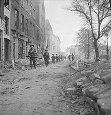 A line of men slowly advancing towards the camera along a ruined city street. They are heavily armed with weapons and ammunition.